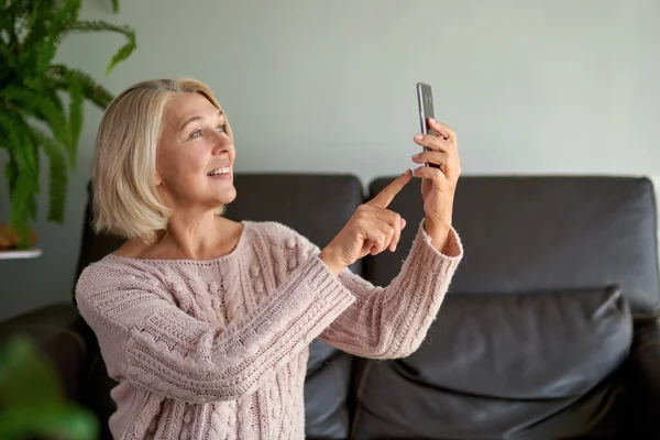 Gelukkige oudere vrouw met behulp van een telefoontje zittend op een bank in de woonkamer in een huis — Stockfoto