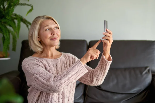 Gelukkige oudere vrouw met behulp van een telefoontje zittend op een bank in de woonkamer in een huis — Stockfoto