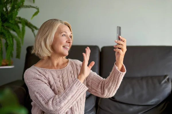 Gelukkige oudere vrouw met behulp van een telefoontje zittend op een bank in de woonkamer in een huis — Stockfoto