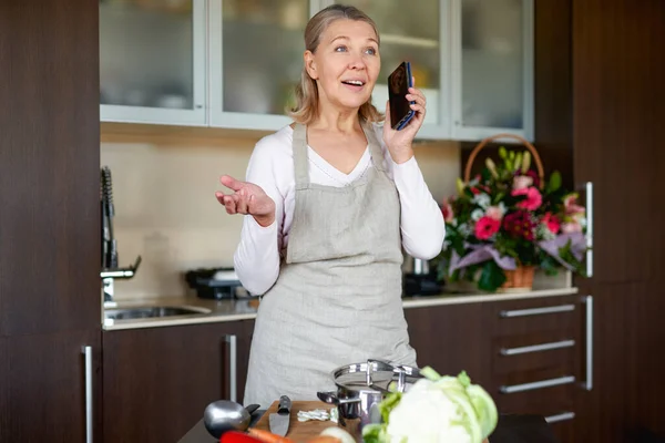 Mature woman in kitchen preparing food and holding smart phone. — Stock Photo, Image