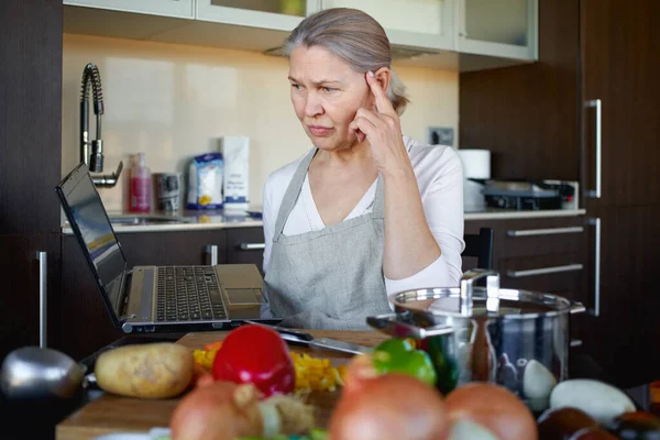 Sad mature woman in kitchen prepares food and looks into laptop