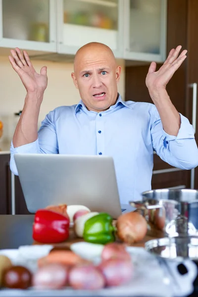 Brutal man in kitchen preparing food and uses laptop.