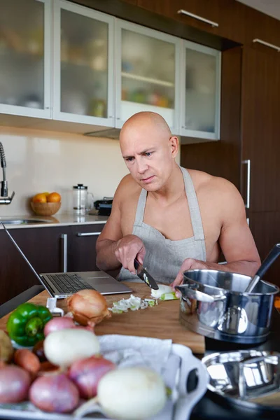 Adult man in kitchen preparing food and looks into laptop. — Stock Photo, Image