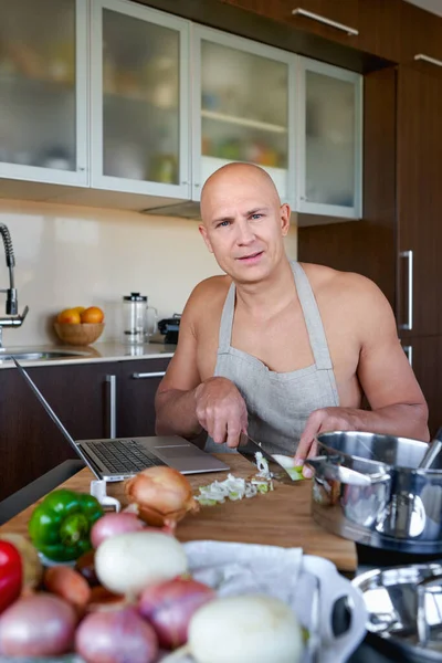 Brutal man in kitchen preparing food and uses laptop. — Stock Photo, Image