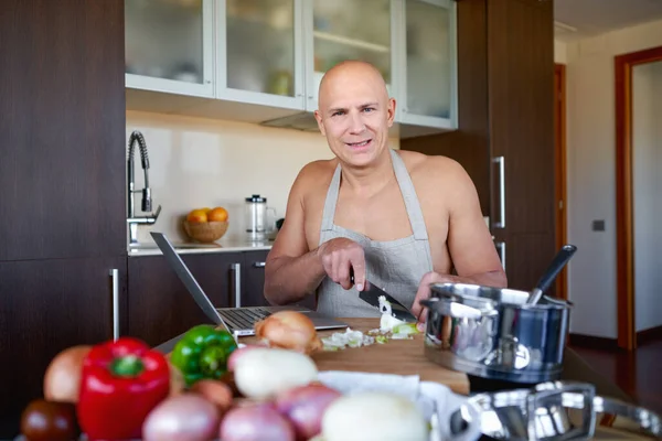 brutal man in kitchen preparing food and uses laptop.