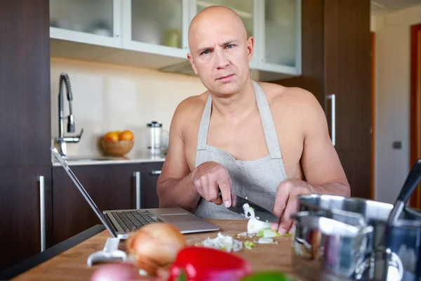 Adult brutal man in kitchen preparing food and looks into laptop.