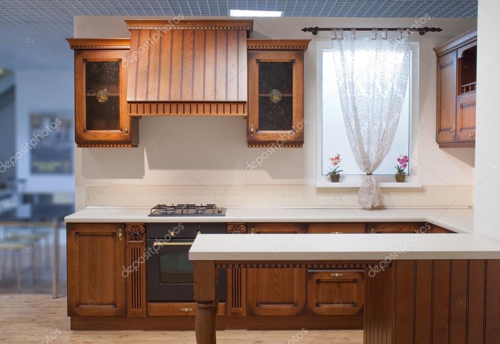 Interior de una cocina con muebles blancos, estantes de pared de madera,  mostradores, horno y ventanas con cortinas, una maqueta