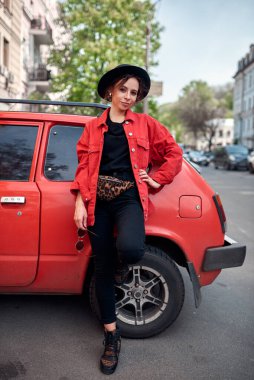 Siyah şapkalı ve kırmızı ceketli bir kız. Caddedeki eski arabanın yanında.