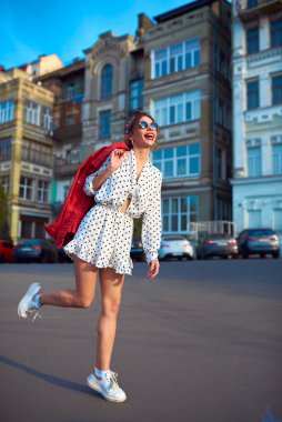Pretty girl in sunglasses and a white short dress rejoices in the sun in the city