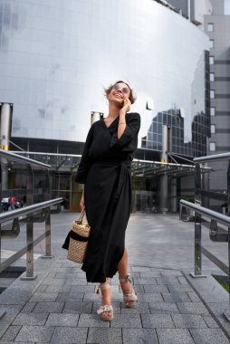 Thin and cheerful woman in black kimono walks down the street and talking on the phone against the background of the modern building
