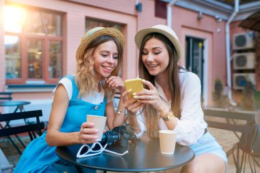 Şapkalı iki tatlı kız arkadaş telefon ekranına bakar ve açık bir kafedeki masada otururken gülümser.