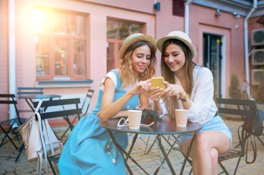 İki tatlı ve neşeli kız bir akıllı telefonun ekranına bakıyor ve açık bir kafede otururken gülüyorlar..