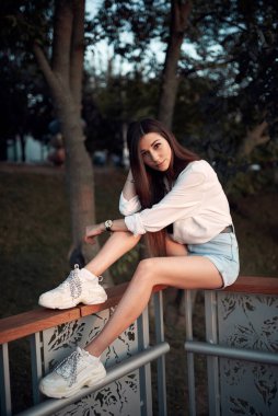 Güzel, genç bir esmer parktaki korkuluklara oturur.