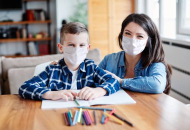 mother teaching son and helping him with homework at home, both of them wearing protective masks