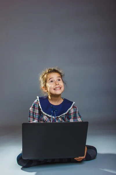 Menina criança sentada jogando laptop surpreso em um fundo cinza — Fotografia de Stock