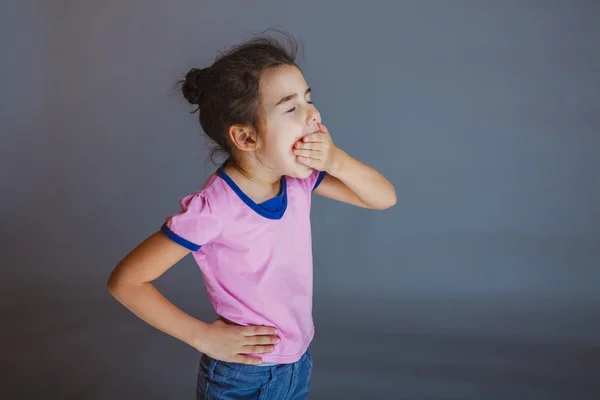 Маленька дівчинка позіхає закриває рот на сірому фоні — стокове фото