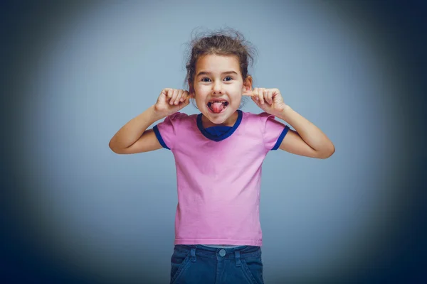 Child girl showing tongue pulls ears teases on a gray background — Stock Photo, Image