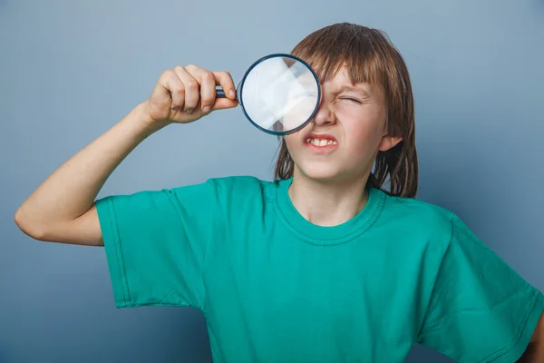 European-looking boy of ten years holding a magnifying glass, a keen eye on gray background — Stock Photo, Image