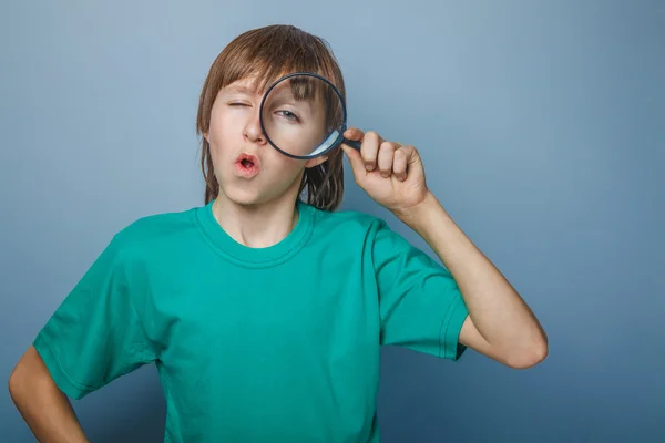 European-looking boy of ten  years holding a magnifying glass, a keen eye on gray background — Stock Photo, Image