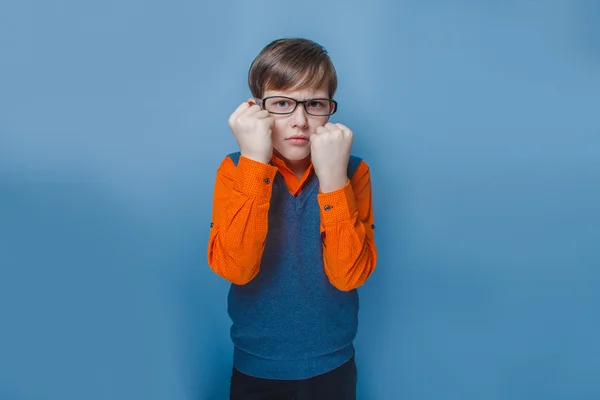 Chico de aspecto europeo de diez años en gafas frunciendo el ceño, muestra los puños infelices sobre un fondo azul —  Fotos de Stock