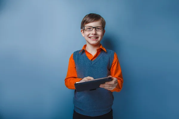 Chico de aspecto europeo de diez años en gafas sosteniendo la tableta en la mano, juega sobre un fondo azul —  Fotos de Stock