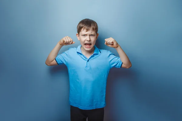 Ragazzo adolescente aspetto europeo in blu T-shirt mostra il potere delle mani su uno sfondo grigio, urlando, muscoli — Foto Stock