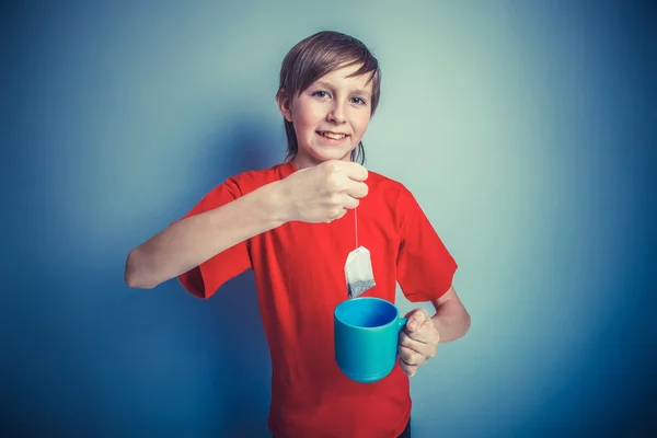 European-looking boy of twelve mug in hand, tea bag on a gray ba — Stock Photo, Image