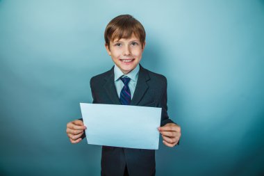European appearance teenager boy with blond hair in suit and tie holding a sheet of white paper on a gray background, smile