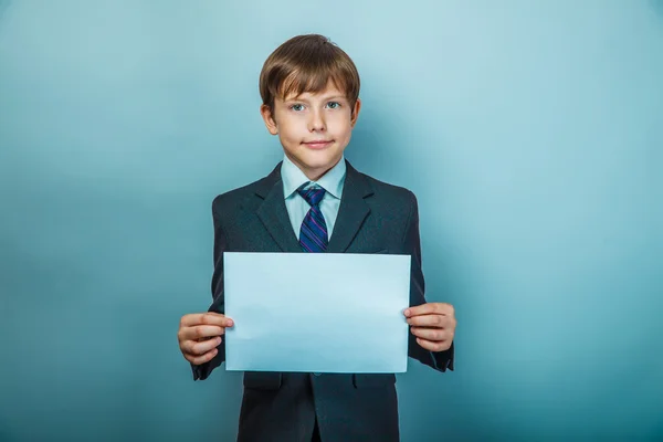 Teen boy of European appearance with brown hair in a suit and tie holding a sheet of white paper on gray background, courtesy — Zdjęcie stockowe