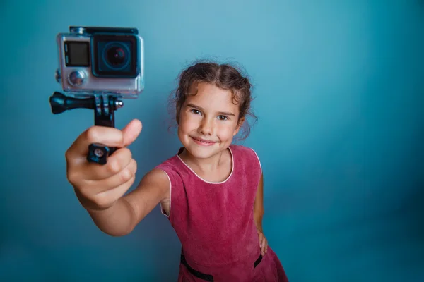 Teen girl child seven years, European appearance brunette holding action camera and smiling on a gray background, shot, joy — Stok fotoğraf