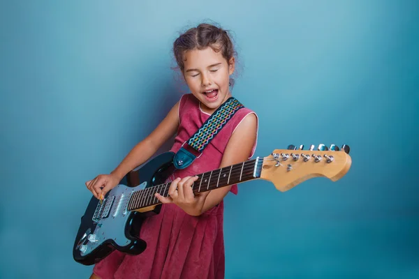 Ragazza adolescente bambino sette anni, aspetto europeo bruna in un abito rosa suona una chitarra e canta i suoi occhi chiusi su uno sfondo grigio, musica, canzone — Foto Stock