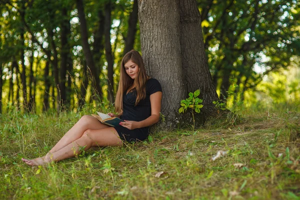 Woman girl sits reading a book under a tree in the forest park o — Stockfoto