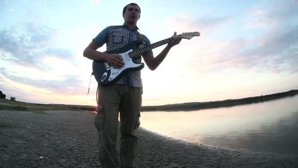 Guitarrista músico homem começa a tocar guitarra elétrica filtro amarelo no rio ao pôr do sol céu nuvens reflexão na água Vídeo — Vídeo de Stock