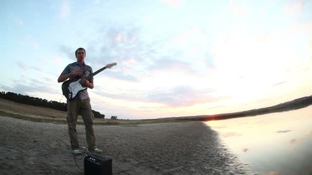 Guitarrista músico hombre deja de tocar la guitarra eléctrica en el río y sale al atardecer cielo nubes reflejo en el agua Video — Vídeos de Stock