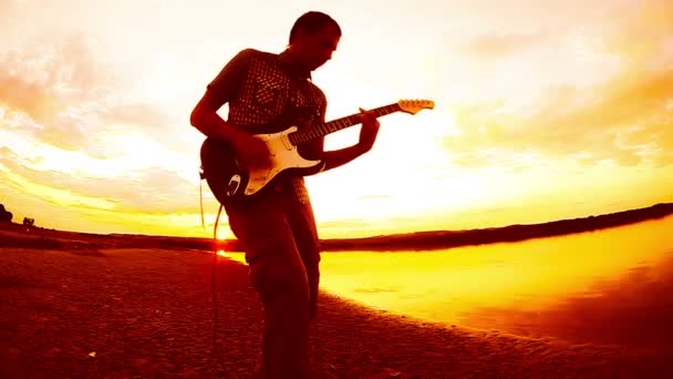 Гитарист оранжевого цвета человек играет соло на электрогитаре у реки на закате неба облака — стоковое видео