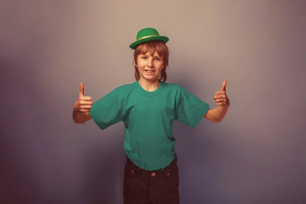 European-looking boy of ten years thumbs up in a hat on a gray b — Stock Photo, Image