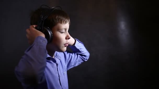 Tonårspojke i en blå skjorta i stora hörlurar lyssna musik mörk bakgrund video — Stockvideo