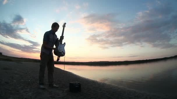 太陽が沈む水によって日没でエレキギターを弾いて男性ギタリスト男ストリートミュージ シャン — ストック動画