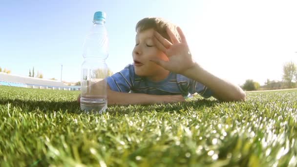 Adolescente bebiendo agua de niño de una botella de plástico que yace en la hierba sonriendo felices veranos de infancia — Vídeos de Stock