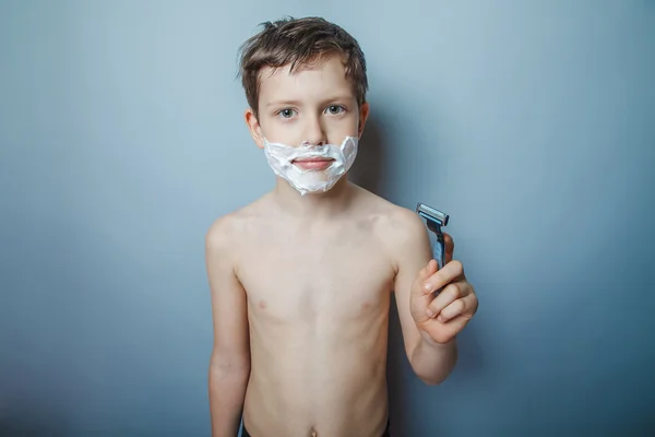 Boy  teenager European appearance  shaves face on a gray — Stockfoto