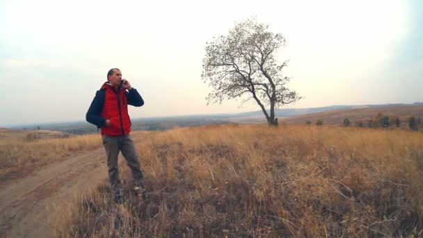 Homem viajante falando sobre a natureza do telefone árvore solitária no outono em uma viagem casaco vermelho — Vídeo de Stock