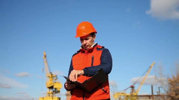 Hombre trabajador de la construcción en un casco naranja que sostiene documentos en el cielo azul de fondo y una grúa — Vídeo de stock