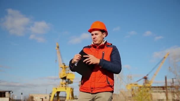 Bauarbeiter mit Helm korrigiert orangefarbene Schreie gegen den blauen Himmel und den Kran — Stockvideo