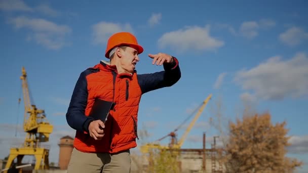 Homme constructeur travailleur de la construction dans un casque orange agitant appel cours derrière la grue et le ciel bleu — Video