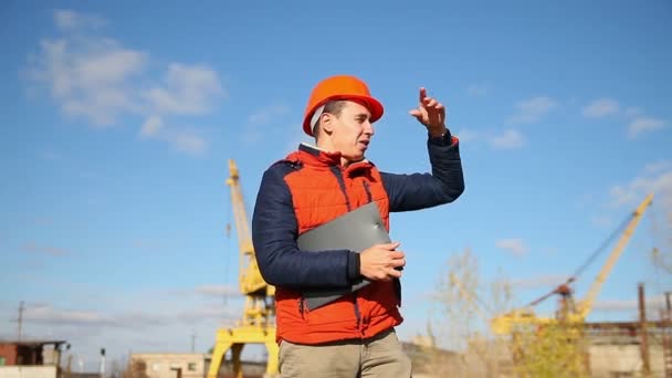Travailleur de la construction masculin dans un casque orange agitant appel cours derrière la grue et le ciel bleu — Video