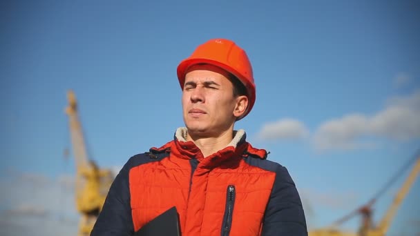 Портрет человека строителя в оранжевом шлеме на фоне голубого неба и строительства — стоковое видео