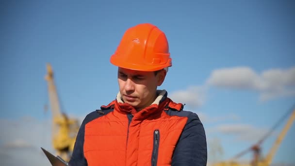 Porträtt av en man byggmästare i orange hjälm ser instrument mot den blå himlen och byggnad — Stockvideo