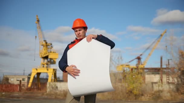 Portrait of man builder in orange helmet looks instruments project against the blue sky and building — Stock Video