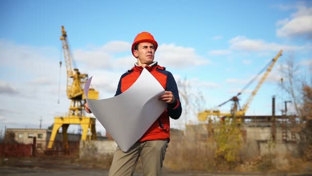 Portrait homme constructeur en casque orange regarde projet d'instruments contre le ciel bleu et la construction — Video