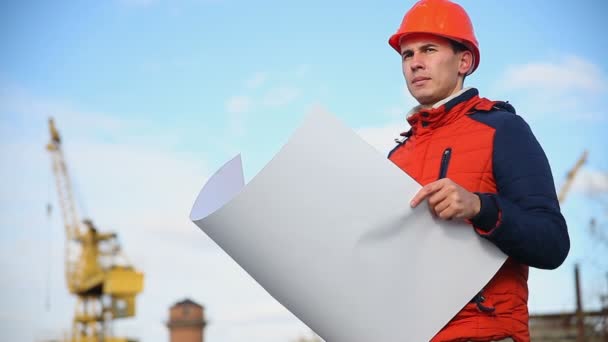 Man portret architect bouwer in oranje helm ziet er instrumenten project tegen blauwe hemel en gebouw — Stockvideo